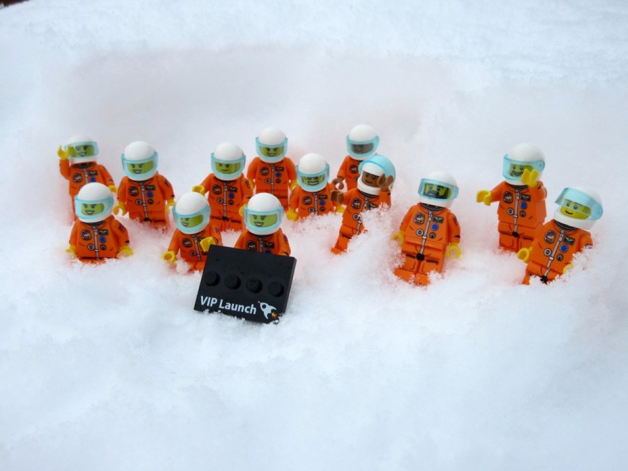 Thirteen Lego astronauts in orange jumpsuits nestled in the snow holding a black sign that reads 'VIP Launch'